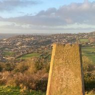 View from the top of Solsbury Hill, lit with low sun. In the foreground is a large stone marker. The city of Bath is visible in the background.