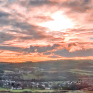 A view from high above a valley looks out over a meandering river, woodlands and hills with the low winter sun shining through streaks of high cloud