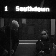 a black and white photo through the side window of a bus waiting at the Bath bus station. A woman seated on the bus looks directly at you while an elderley man is sitting himself down beside her. The glow of the bus's route sign is overexposed and hazy and reflected in the dark windshielf of a second bus parked next to it, off to the side and a little nearer to the viewer.