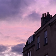 The upper floors of two Georgian buildings in Bath, viewed from below. Behind them is the sky just before sunrise, in shades of pink and purple, with some patchy clouds and a seagull flying past. One of the buildings has some Christmas lights.