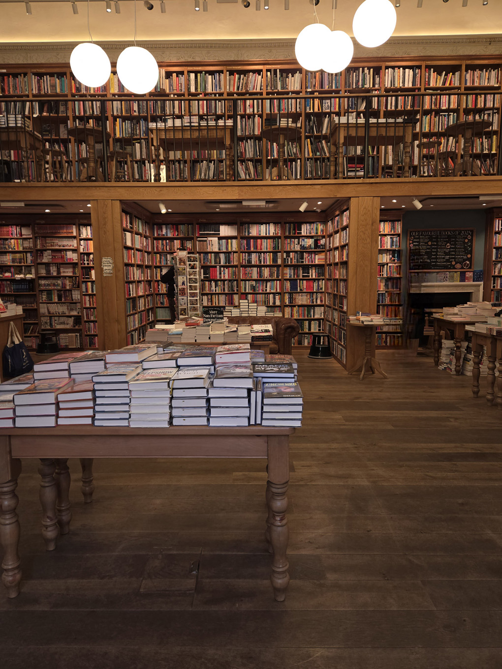 Inside a bookstore. There is a table stacked with books in the centre and all the walls are stacked floor to ceiling with books. Against a wall, there is a wooden ladder on a rail that runs the length of the shelves