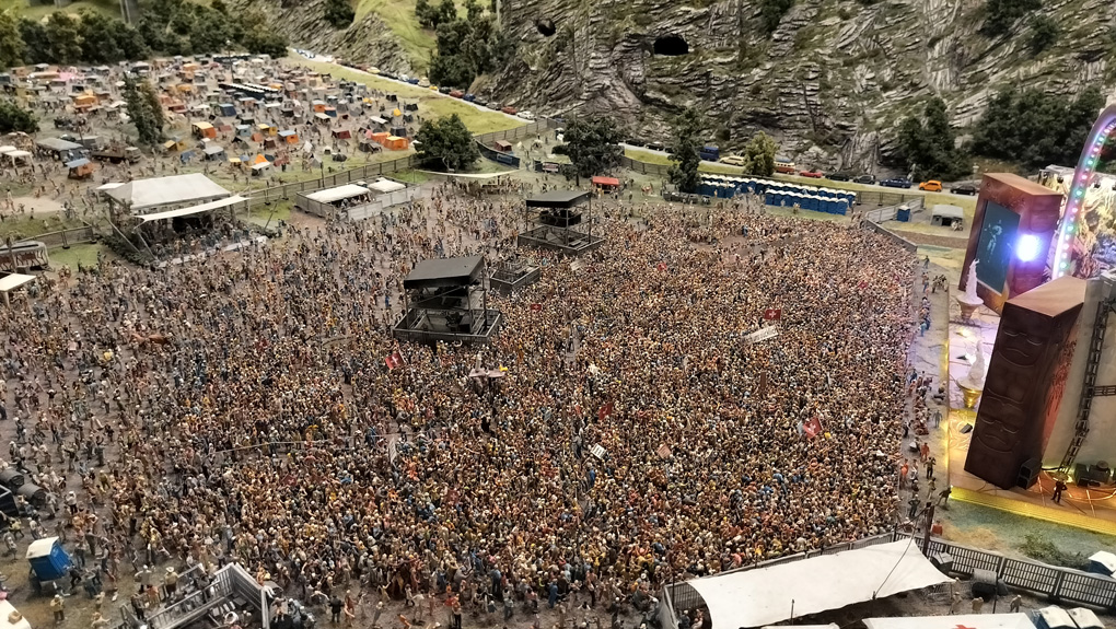 To the casual observer a photo of a busy festival site, taken from an elevated point, surveying the whole scene. The main stage is on the right, there is a large crowd and, in the distance, a car park. The area is surrounded by mountainous terrain.