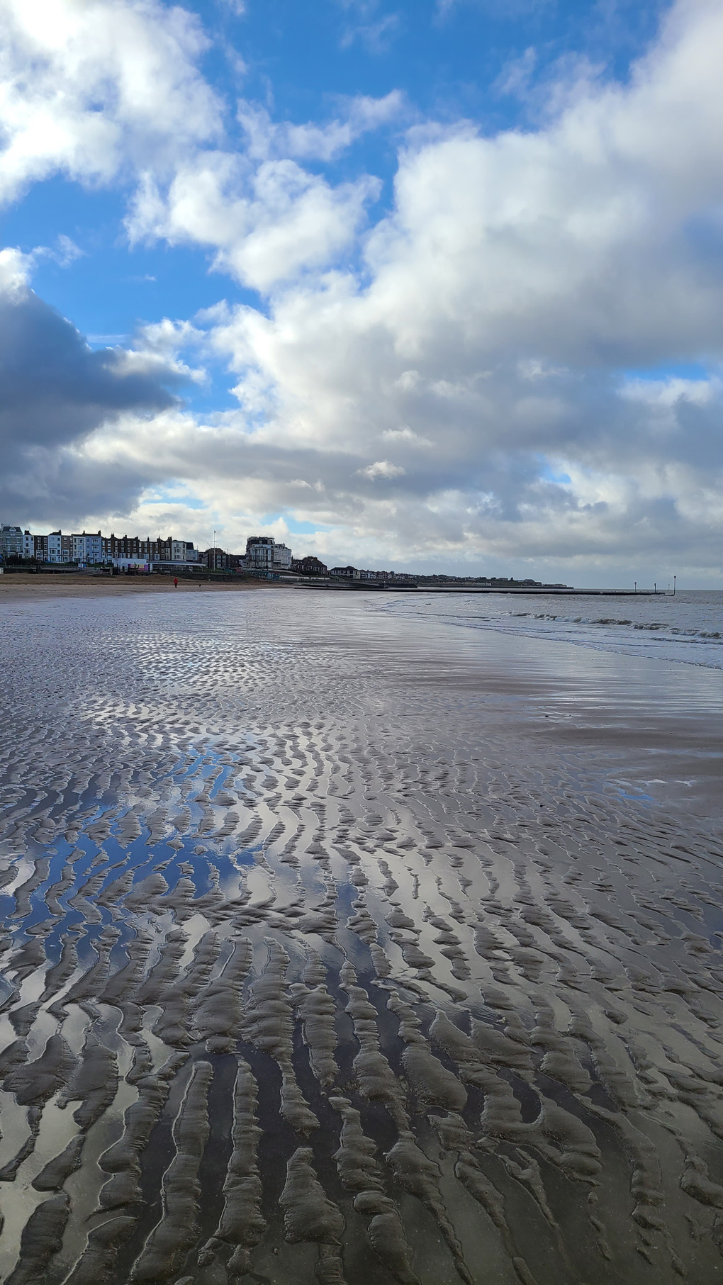 The top half shows a bright blue cloudy sky. The bottom half shows that sky reflected in the water pooled in the rippled sand of the beach. The horizon in the centre shows Margate, a variety of buildings.