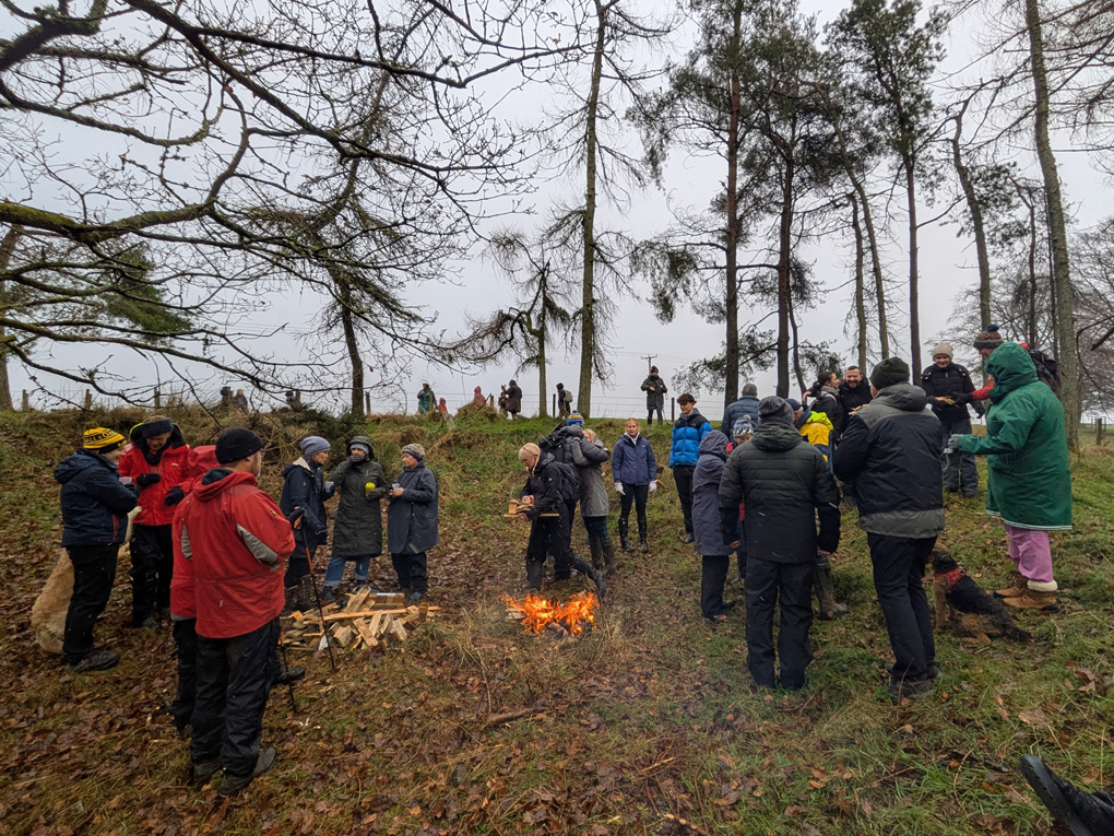 Numerous warmly attired villagers gathered around a bonfire in the cold woods with a dram of whisky for a neighbourly blether