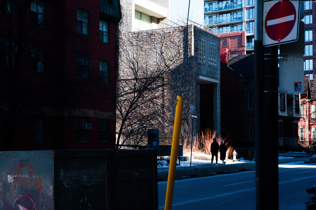 Couple walks down a quiet street in downtown Toronto on a bright day after snowfall. They are silhouetted in a bright patch of sun and light reflected off snow on the ground