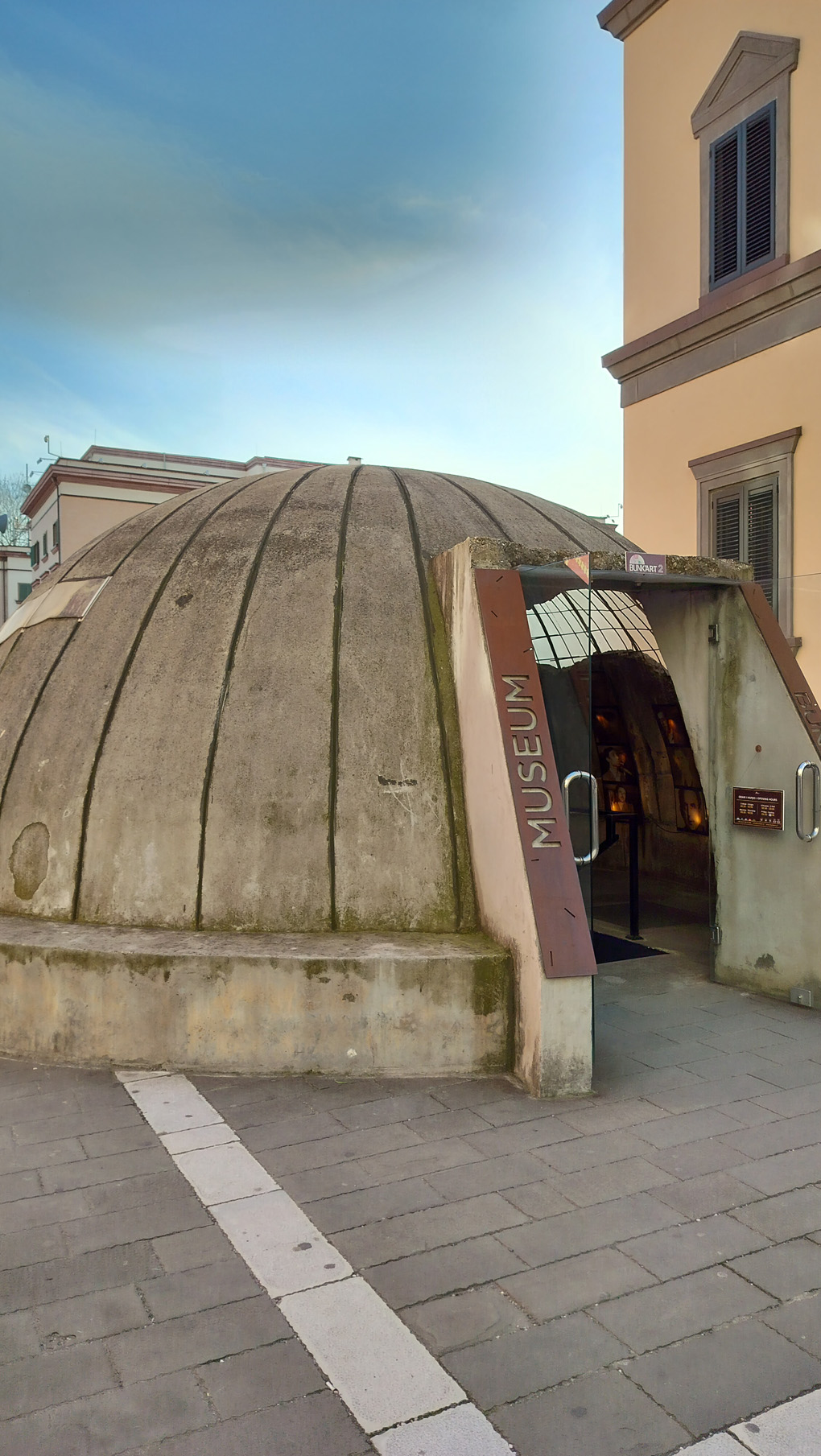 On a pedestrianized street, outside a yellow building, there is a concrete dome, about 10 foot tall, with a door at the front an the words 