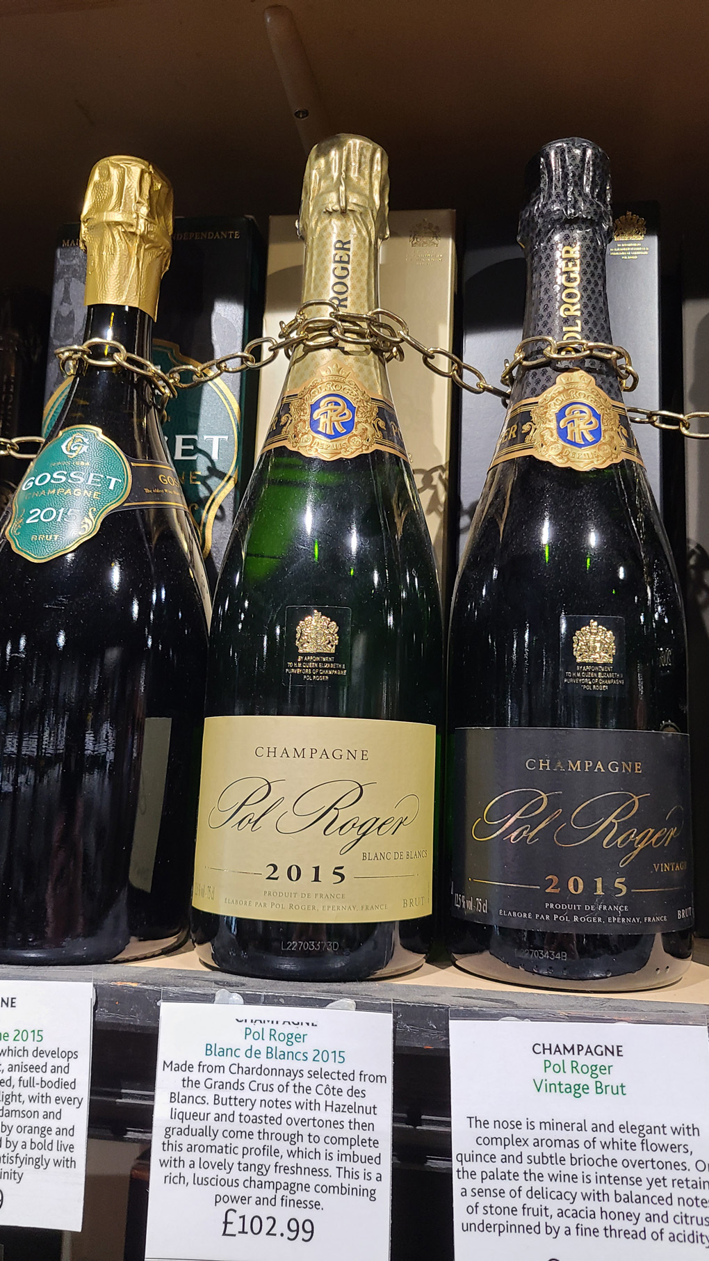 3 bottles of Champagne on a shelf with a chain wrapped around the necks to connect them. Below them are white squares of card with descriptions of each Champagne.