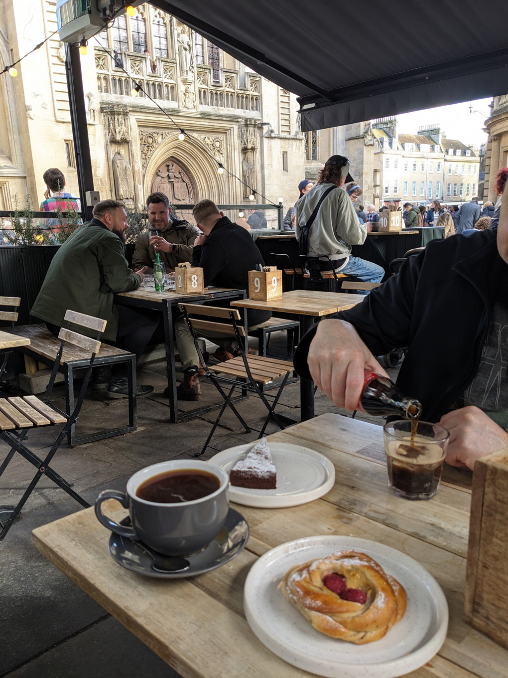 View from a table outside at a cafe next to Bath Abbey. The sun is shining, there is a plate with pastries on the table and beyond the tables there is the front of the Abbey.