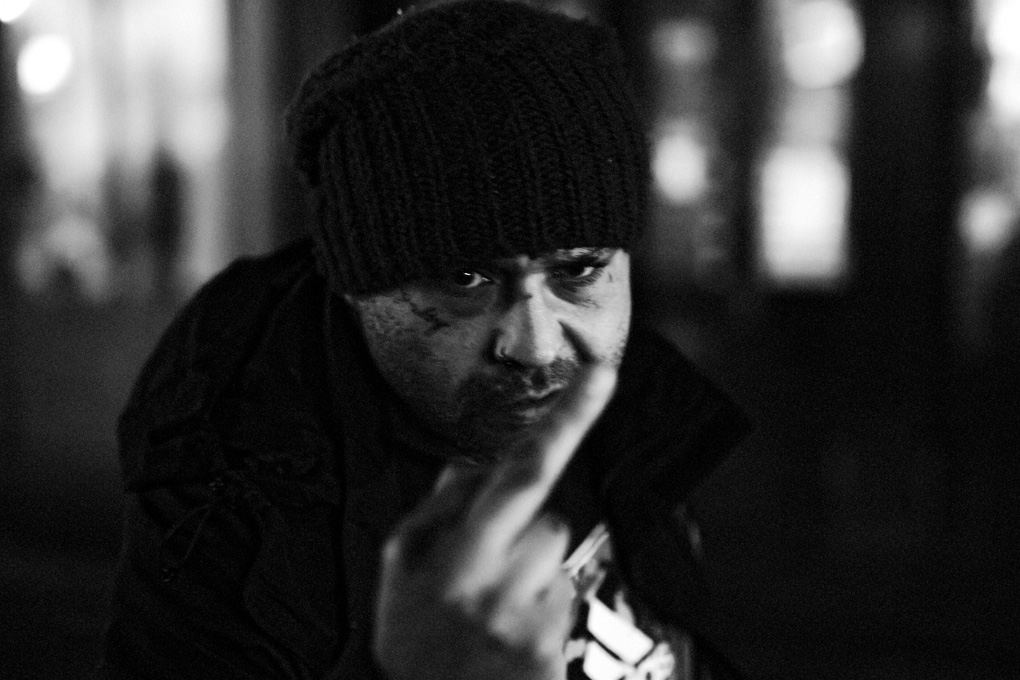 A man angrily flipping off the photographer, in black and white. He is wearing a knit beanie hat, has a small tattoo under his right eye, and is glaring at close range directly into the camera. His finger covers the lower part of his face.