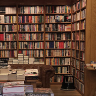 Inside a bookstore. There is a table stacked with books in the centre and all the walls are stacked floor to ceiling with books. Against a wall, there is a wooden ladder on a rail that runs the length of the shelves