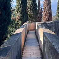 A path in the sunshine bordered by two castle walls, beyond which on each side is a sharp drop. The walls have trees growing on each side.