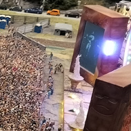 To the casual observer a photo of a busy festival site, taken from an elevated point, surveying the whole scene. The main stage is on the right, there is a large crowd and, in the distance, a car park. The area is surrounded by mountainous terrain.