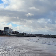 The top half shows a bright blue cloudy sky. The bottom half shows that sky reflected in the water pooled in the rippled sand of the beach. The horizon in the centre shows Margate, a variety of buildings.