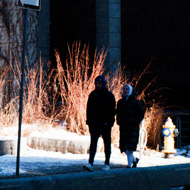 Couple walks down a quiet street in downtown Toronto on a bright day after snowfall. They are silhouetted in a bright patch of sun and light reflected off snow on the ground