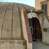 On a pedestrianized street, outside a yellow building, there is a concrete dome, about 10 foot tall, with a door at the front an the words 