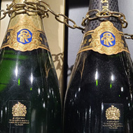 3 bottles of Champagne on a shelf with a chain wrapped around the necks to connect them. Below them are white squares of card with descriptions of each Champagne.