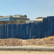 Two women walk in front of a tall blue corrugated fence which runs along a dusty roadside in the bright African sun behind it new buildings are being constructed