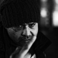 A man angrily flipping off the photographer, in black and white. He is wearing a knit beanie hat, has a small tattoo under his right eye, and is glaring at close range directly into the camera. His finger covers the lower part of his face.